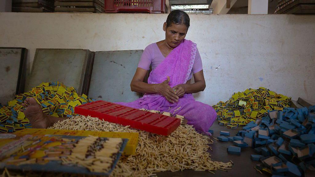 Mulher t&acirc;mil de s&aacute;ri rosa sentada no ch&atilde;o, embalando centenas de caixas de f&oacute;sforos em grandes pilhas