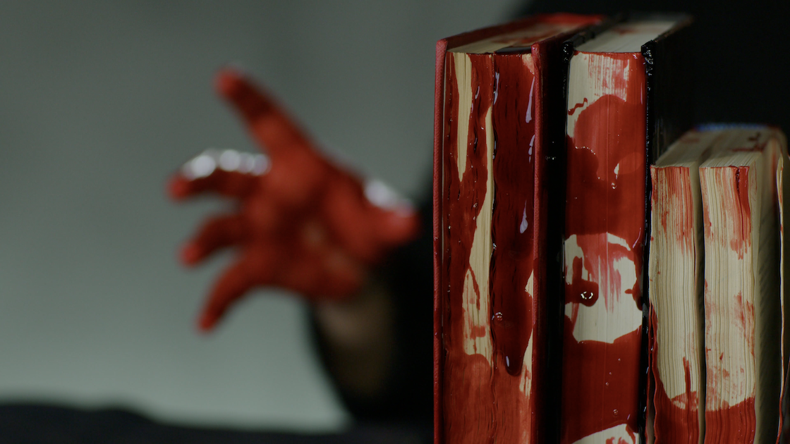 في المقدّمة، تظهر ثلاثة كتب مصطفّة تتلطّخ أغلفتها بالدم، وفي الخلفية تمدّ يدٌ مغطّاة بالدماء نفسها إلى الأمام، على خلفية رمادية اللون.