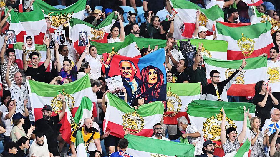 Los aficionados iraníes en las gradas ondean banderas verdes, blancas y rojas, con un escudo dorado en la mitad.
