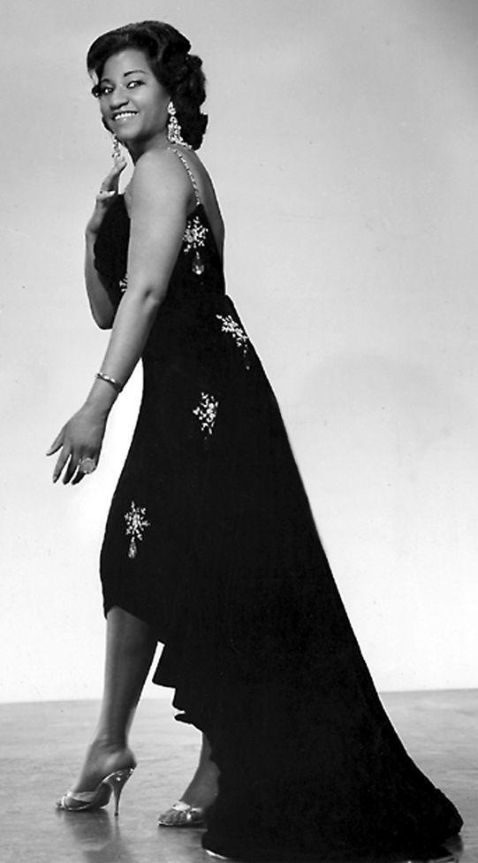 Celia Cruz, con un elegante vestido negro y tacones, de pie