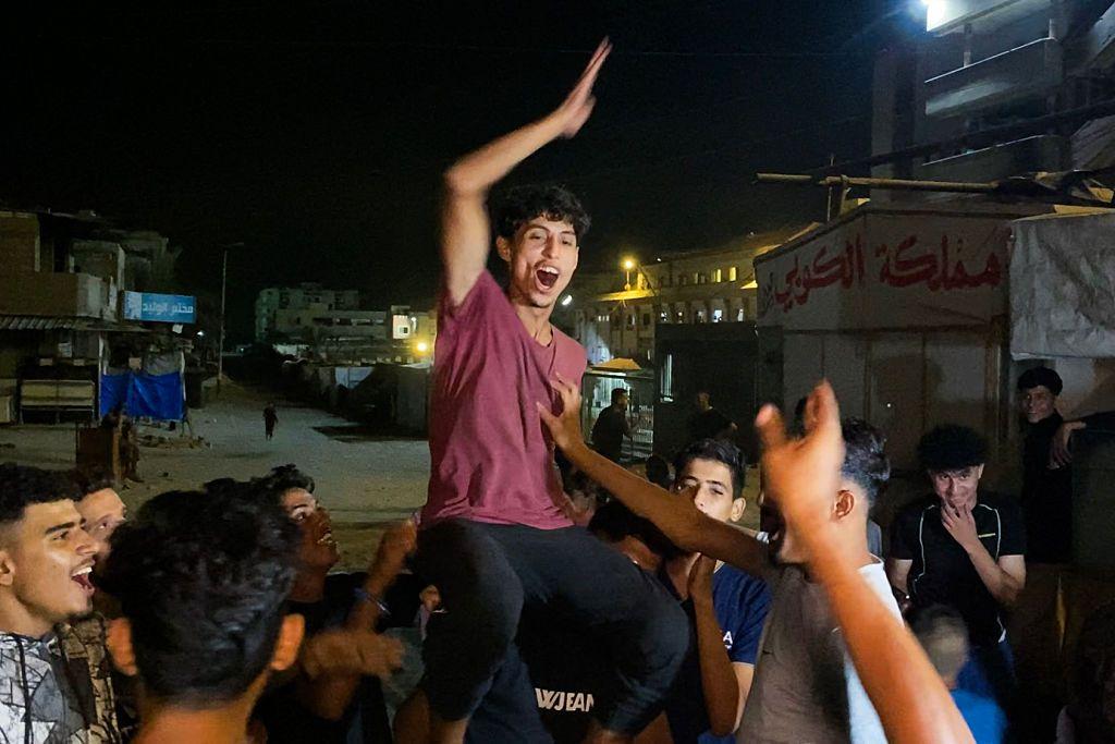 Un grupo de hombres jóvenes palestinos están en círculo con caras de júbilo y sostienen a uno de ellos elevado, sobre los hombros de uno otro. El que está elevado tiene su brazo levantado, con una gran expresión de júbilo. Todos visten ropa casual, camisetas y pantalón deportivo. Es de noche.