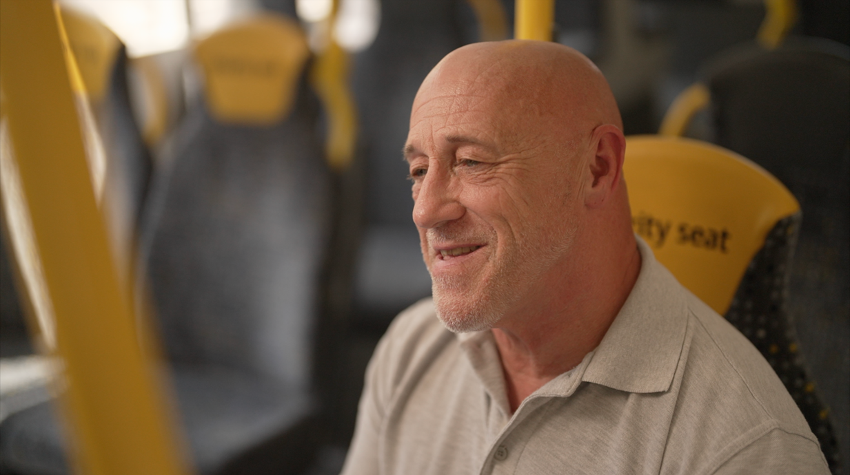 Dave Green, homem careca de meia idade, veste uma camisa polo cinza clara e est&aacute; sentado em uma poltrona de &ocirc;nibus