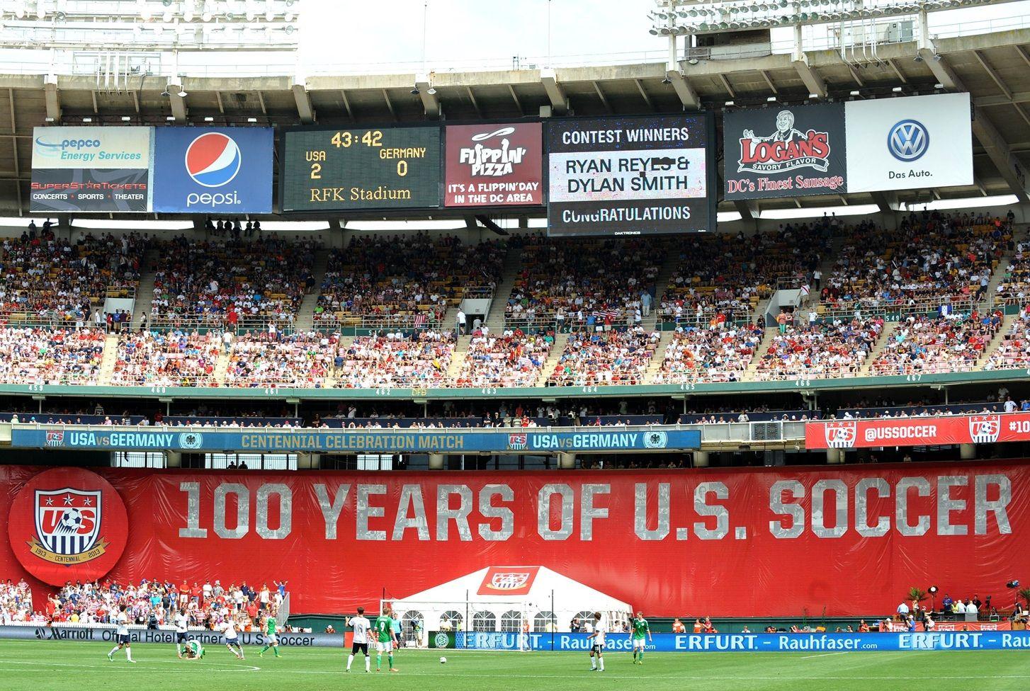 Los jugadores juegan en la cancha. Se ven las gradas del estadio y una pancarta gigante de fondo rojo que dice 100 years of U.S. Soccer. Hay una pantalla electrónica que muestra el tiempo y el marcador: 2 para USA y 0 para Alemania. Se ven publicidades de compañías.
