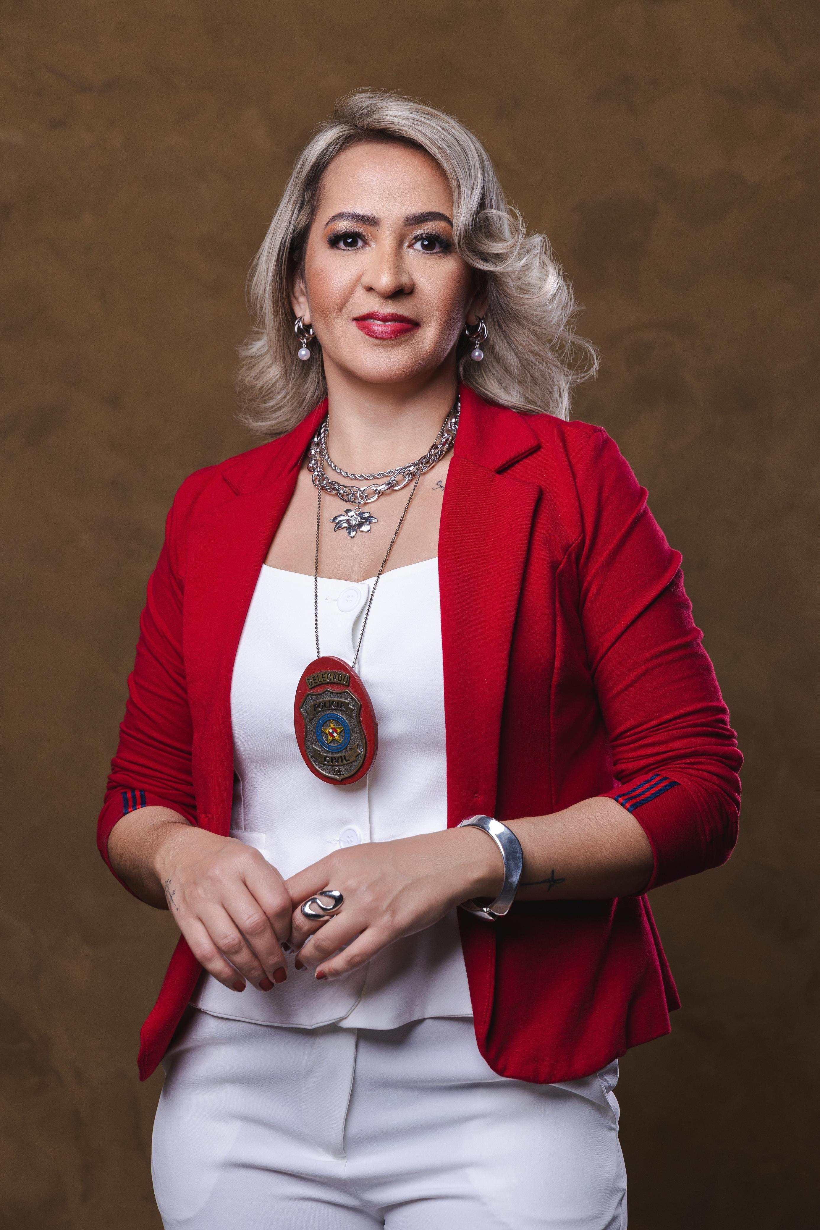 Amanda Souza veste um terninho vermelho e blusa e cal&ccedil;as brancas, usando no pesco&ccedil;o um distintivo da Pol&iacute;cia Civil com o fundo tamb&eacute;m na cor vermelha. Ela tem os cabelos loiros e usa brincos, colar, pulseira e anel em prata