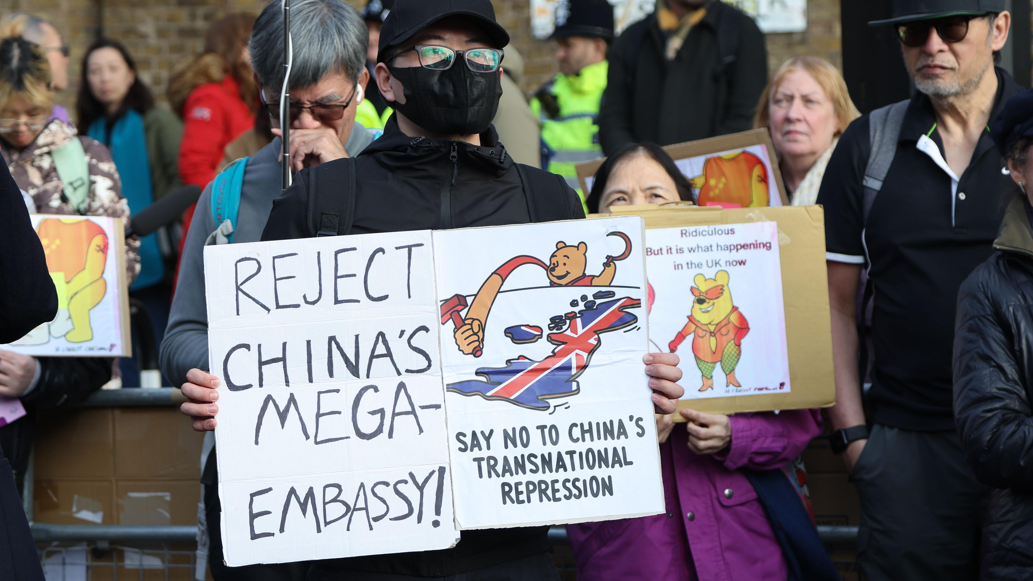 倫敦前皇家鑄幣廠外一名華人示威者舉起反對興建中國「超級大使館」的標語牌，顯示維尼熊伸長手臂，舉起共產黨黨徽上的鐵鎚，碰觸披上英國國旗的英國國土（17/1/2026）