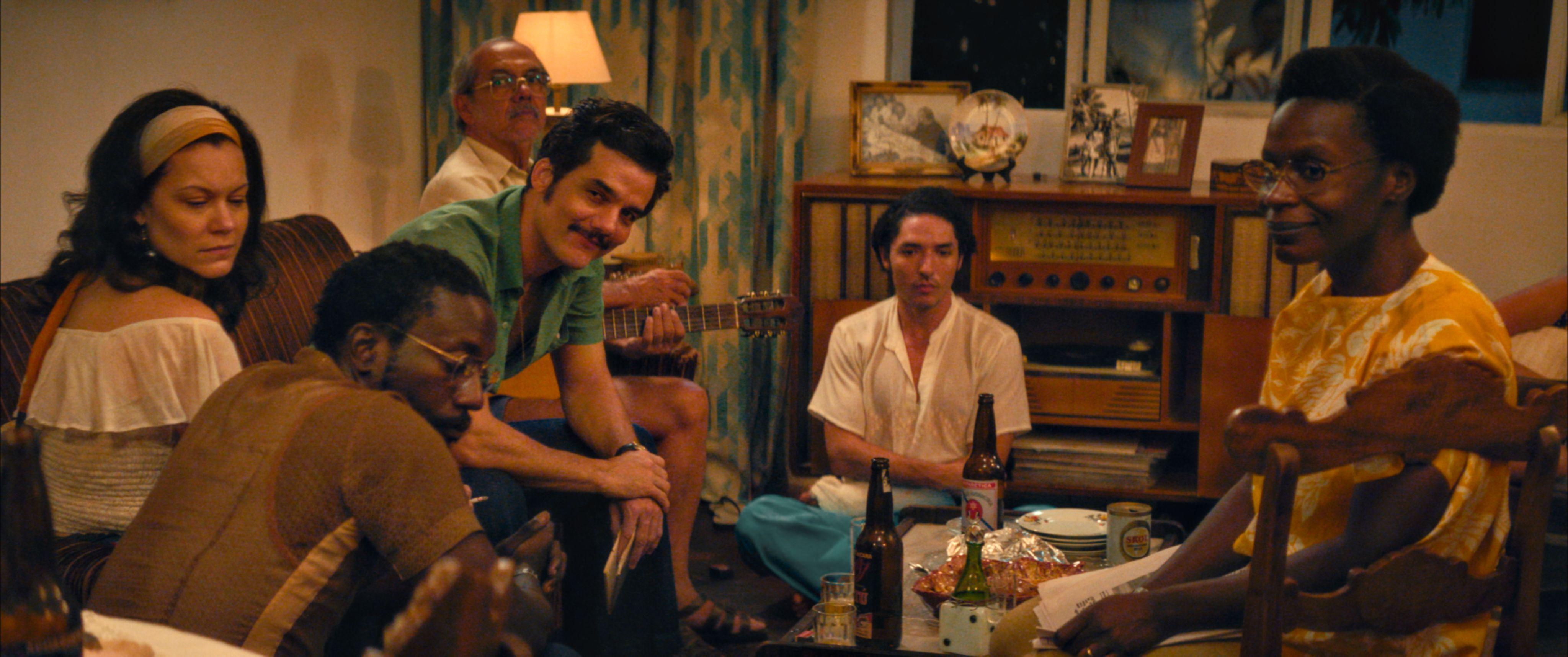 Seis pessoas est&atilde;o reunidas em uma sala de estar, sentadas em volta de uma mesa de centro com garrafas de cerveja, copos e petiscos. Um homem segura um viol&atilde;o enquanto conversa com o grupo. Ao fundo, h&aacute; um r&aacute;dio antigo sobre um m&oacute;vel com fotografias e um abajur aceso. O ambiente tem ilumina&ccedil;&atilde;o quente e decora&ccedil;&atilde;o que lembra uma casa de &eacute;poca, com cortinas estampadas e m&oacute;veis de madeira.