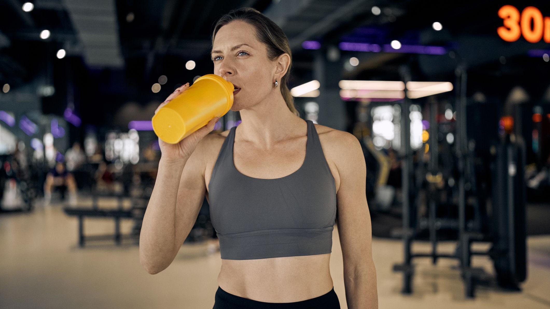 Una mujer bebe de un batidor de proteínas amarillo en un gimnasio. Lleva un sujetador deportivo gris y mira a lo lejos.