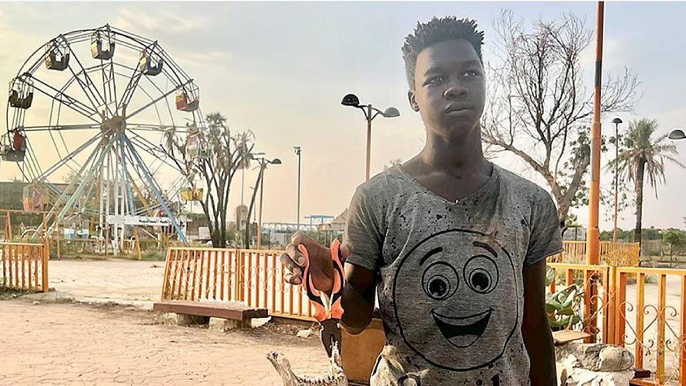 Ahmed en el parque en el que trabaja. Viste una camiseta andrajosa gris que tiene el dibujo de un rostro sonriente y sostiene en su mano con pinzas un trozo de mandíbula humana. Al fondo se ve una rueda gigante vacía.