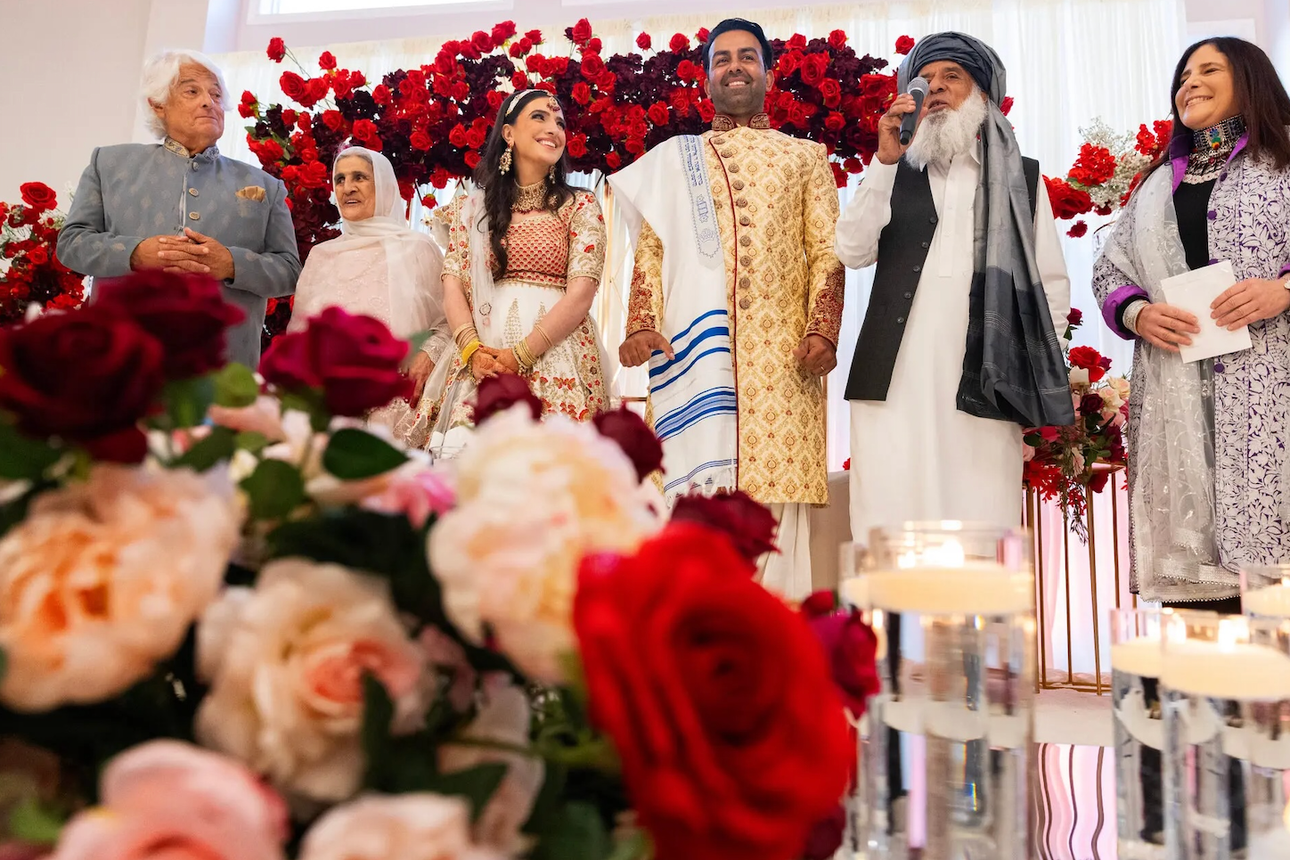 Sammi and Safi standing on the stage with their respective parents during their wedding celebrations. There is a backdrop of red flowers and a blurry close up of colourful flowers. 