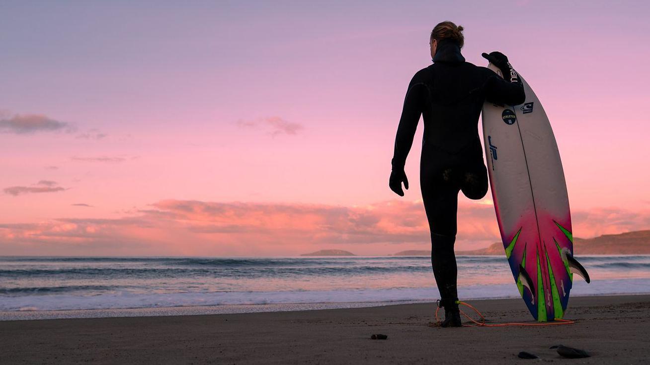 Un surfista con traje de neopreno negro integral y guantes. Está de pie en una playa al atardecer, con una colorida puesta de sol rosa. De espaldas a la cámara, con el mar frente y el vasto cielo al fondo, sostiene una tabla de surf blanca, rosa fucsia y verde, con la punta apuntando hacia el cielo y el otro extremo apoyado en la arena.