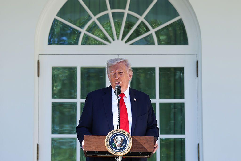 El presidente Donald Trump pronuncia un discurso durante un almuerzo en el Jardín de las Rosas de la Casa Blanca. Va vestido con traje de chaqueta azul oscuro, camisa blanca y corbata roja. Está apoyado en un atril que tiene un micrófono y un escudo de la presidencia de Estados Unidos. El fondo es una pared blanca, en medio una ventana de marcos blancos con una cortina verde.