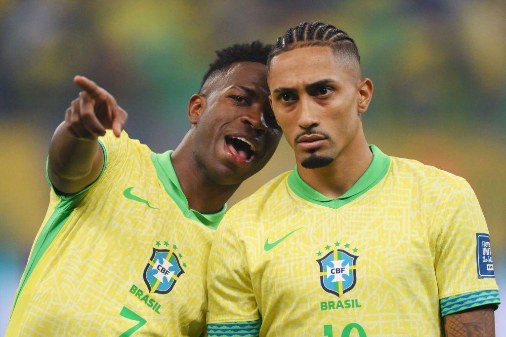 Vinicius Jr. (esq.) e Raphinha (dir.), com a camisa da sele&ccedil;&atilde;o brasileira, conversam no campo de jogo