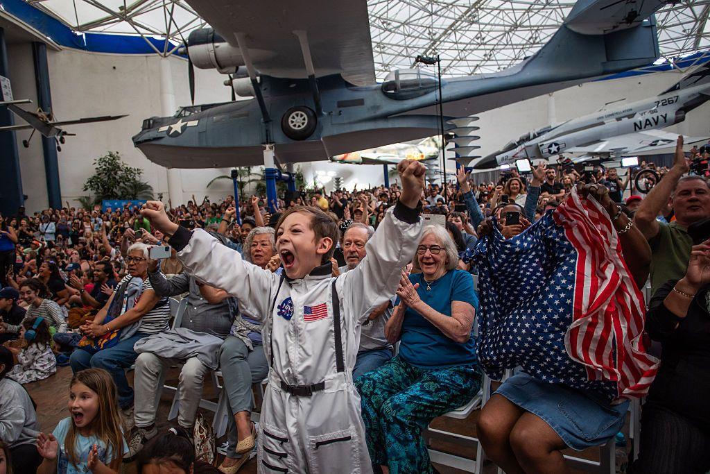Un niño vestido con un disfraz de astronauta anima junto a una mujer que ondea una bandera mientras ven una retransmisión en directo del regreso de los tripulantes del Artemis II a la Tierra en el Museo del Aire y el Espacio de San Diego
