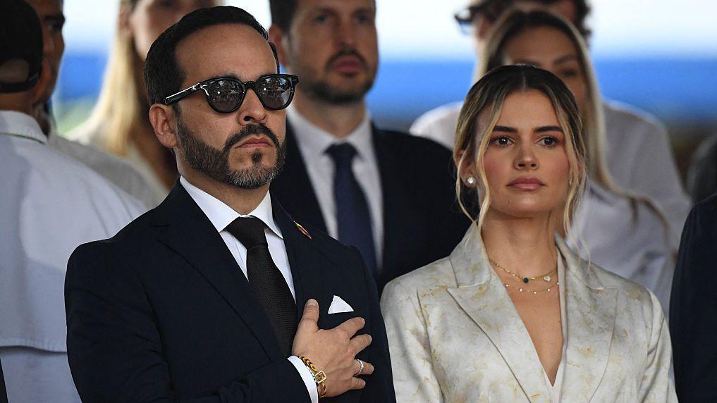 Abelardo de la Espriella, vestido con traje de corbata negro y gafas negras de sol, tiene la mano en el pecho y tiene una mirada sombría. A su lado está Ana Lucia Pineda, vestida con un traje gris. 