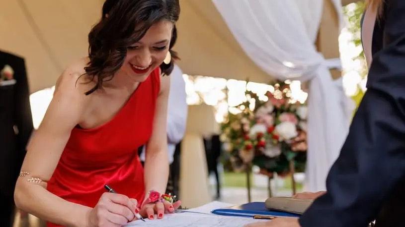 Devojka u crvenoj haljini drži penkalo i potpisuje se u matičnu knjigu venčanih