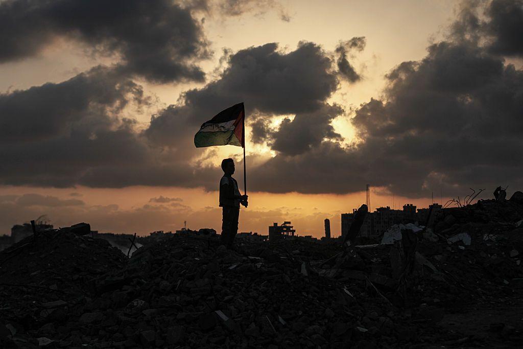 طفل لا تظهر صورته واضحة، يحمل العلم الفلسطيني على مبنى مدمر، السماء فوقه ملبدة بالغيوم والشمس غائبة.