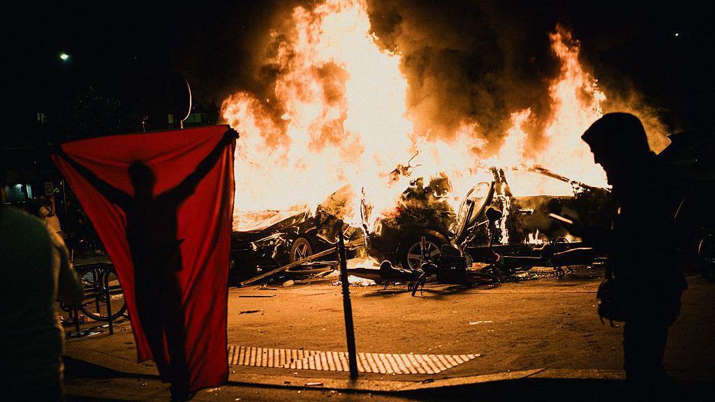 Un coche arde en llamas mientras dos hombres pasan por delante.
