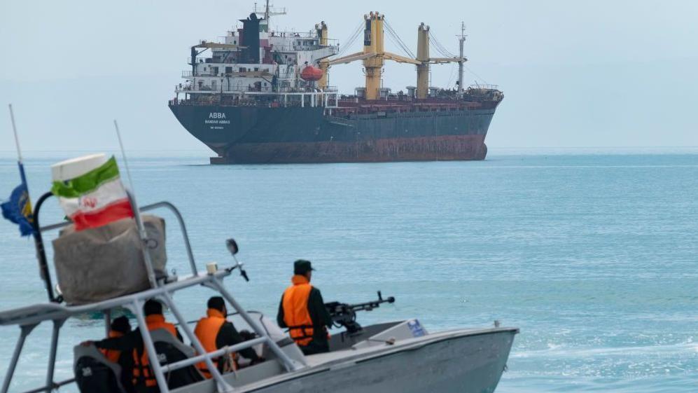 一艘小型巡邏艇在平靜的藍色水面上疾馳，艇上載著幾名身穿亮橙色救生衣的人——其中一人艇首裝有機槍——艇尾飄揚著一面伊朗國旗。遠處，一艘巨大的黑色船隻沉重地停泊在水中，甲板上架著幾台黃色起重機。