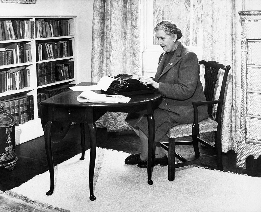 Agatha Christie sentada escribiendo a máquina al lado de su biblioteca.