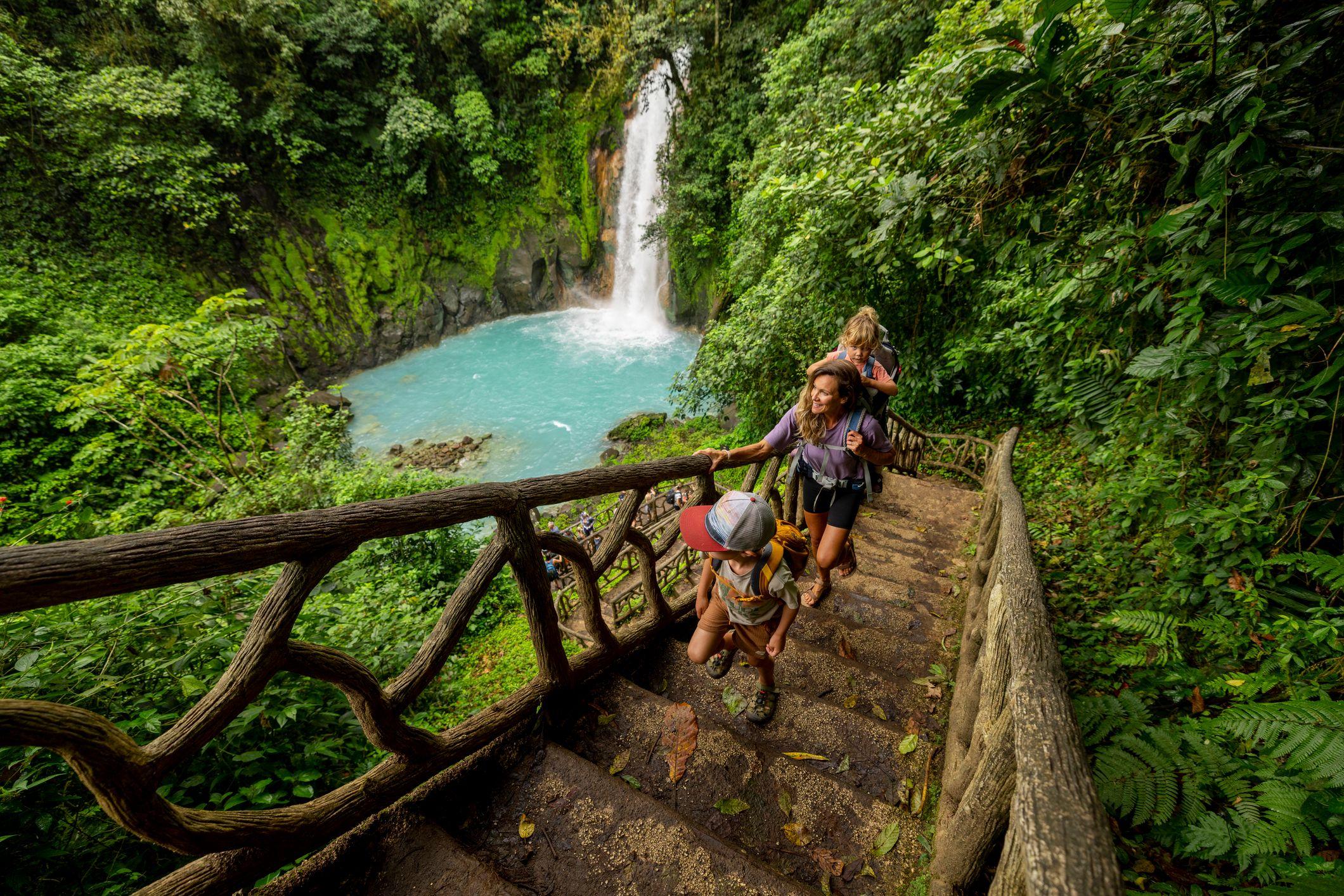 Cascada en Costa Rica rodeada de selva. Una mujer y dos niños caminan por un sendero de madera. 