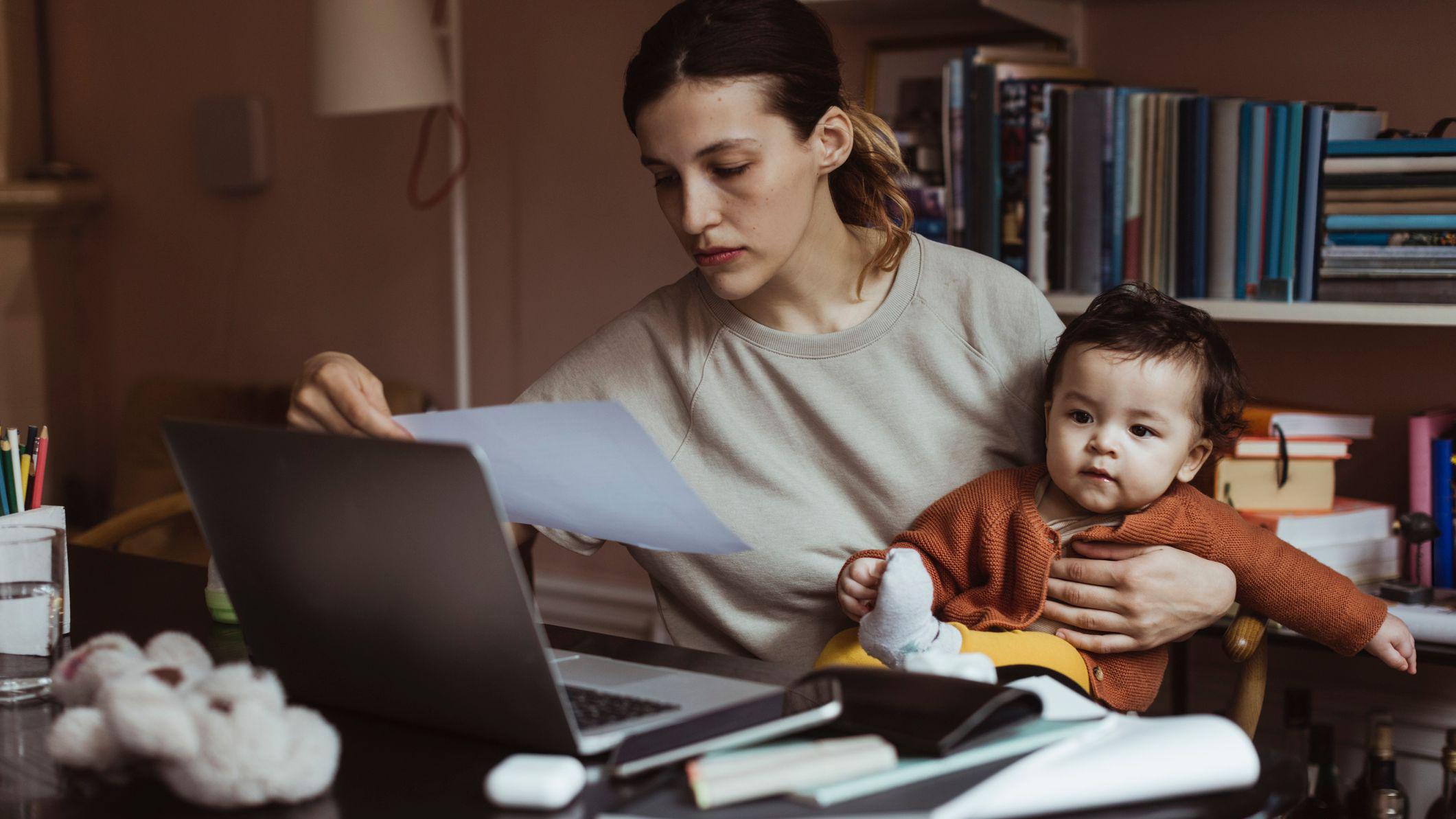 Foto de uma mulher na frente do computador com um bebê no colo. 
