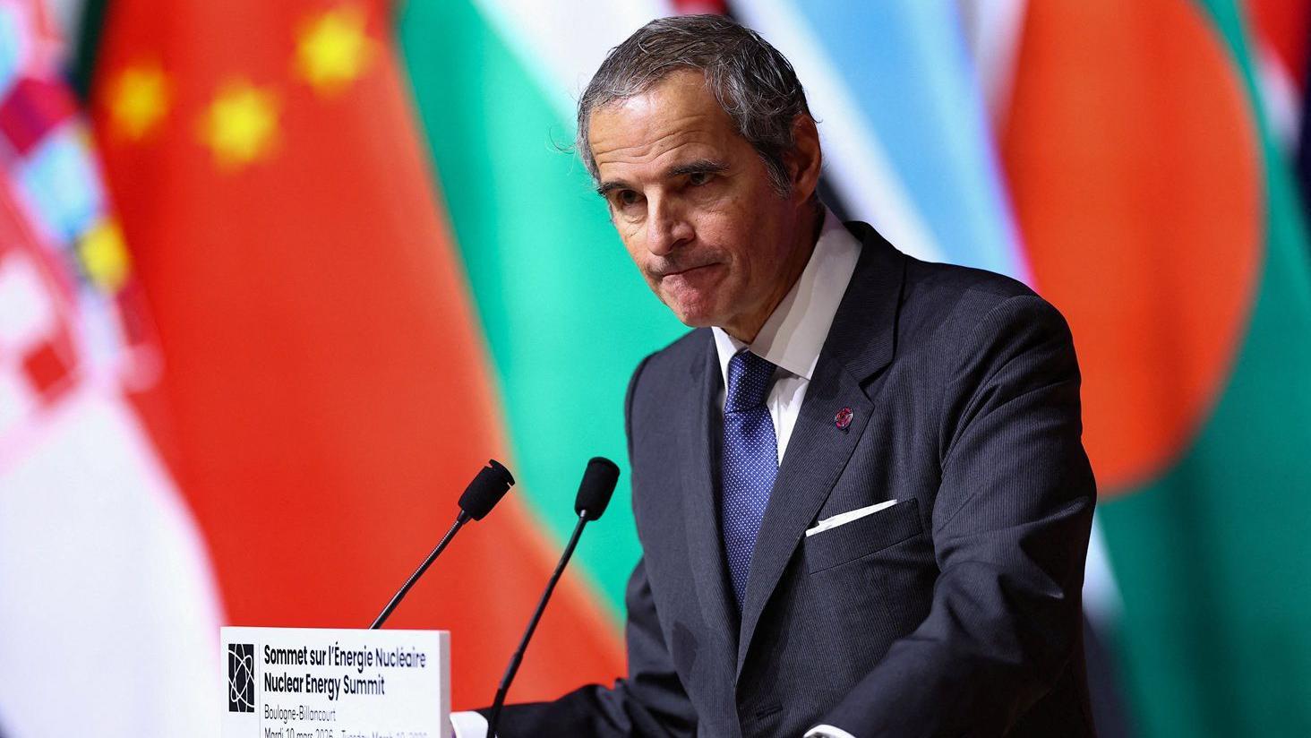 Rafael Grossi en un podio frente a un micrófono y en el fondo se ven varias banderas