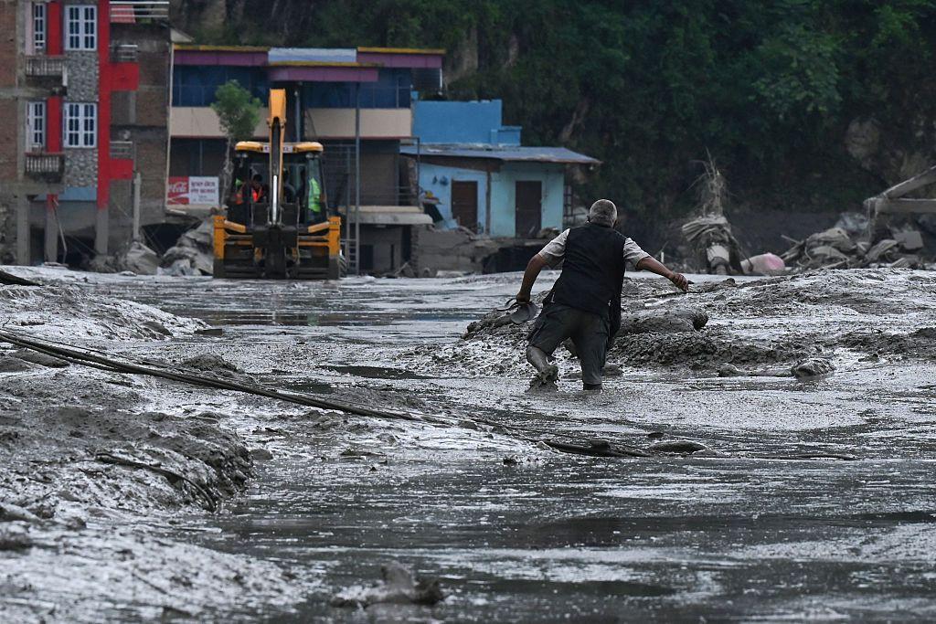 Una persona se abre paso con dificultad entre lodo espeso y gris y agua de inundación poco profunda en una zona urbana gravemente dañada. Al fondo, una excavadora amarilla se encuentra sobre el camino cubierto de lodo. 