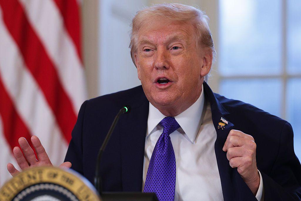 ترامب يرتدي بدلة رسمية تبدو كحلية اللون وقميصاً أبيض وربطة عنق قريبة من اللون البنفسجي منقطة بالكحلي وخلفه العلم الأمريكي. متحدثاً في مؤتمر.