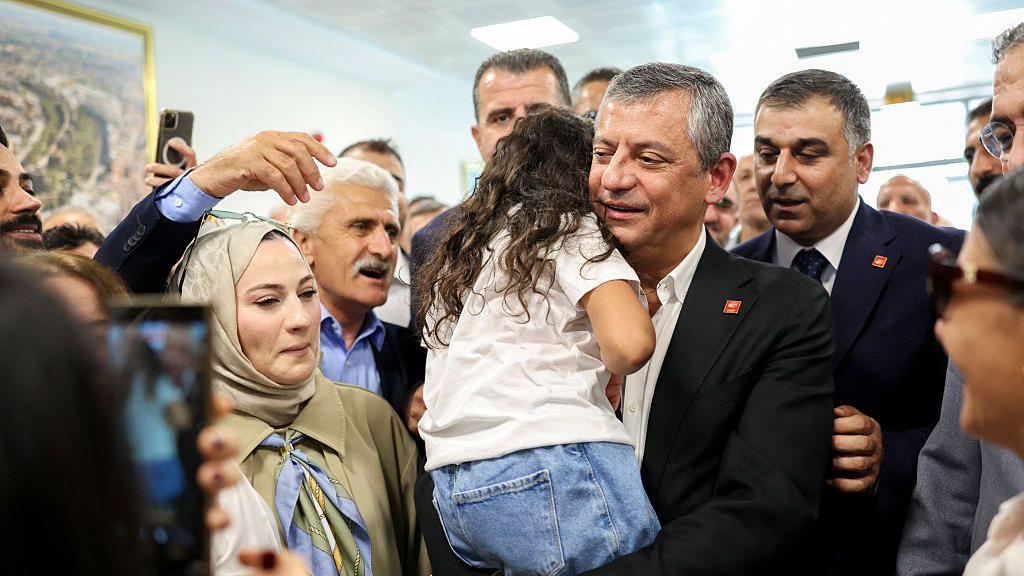 Özgür Özel'in 26 Haziran'da ziyaret ettiği Diyarbakır'da havalimanındaki karşılamadan bir fotoğraf. Kucağında sırtı dönük bir kız çocuk var. etrafı kalabalık. 