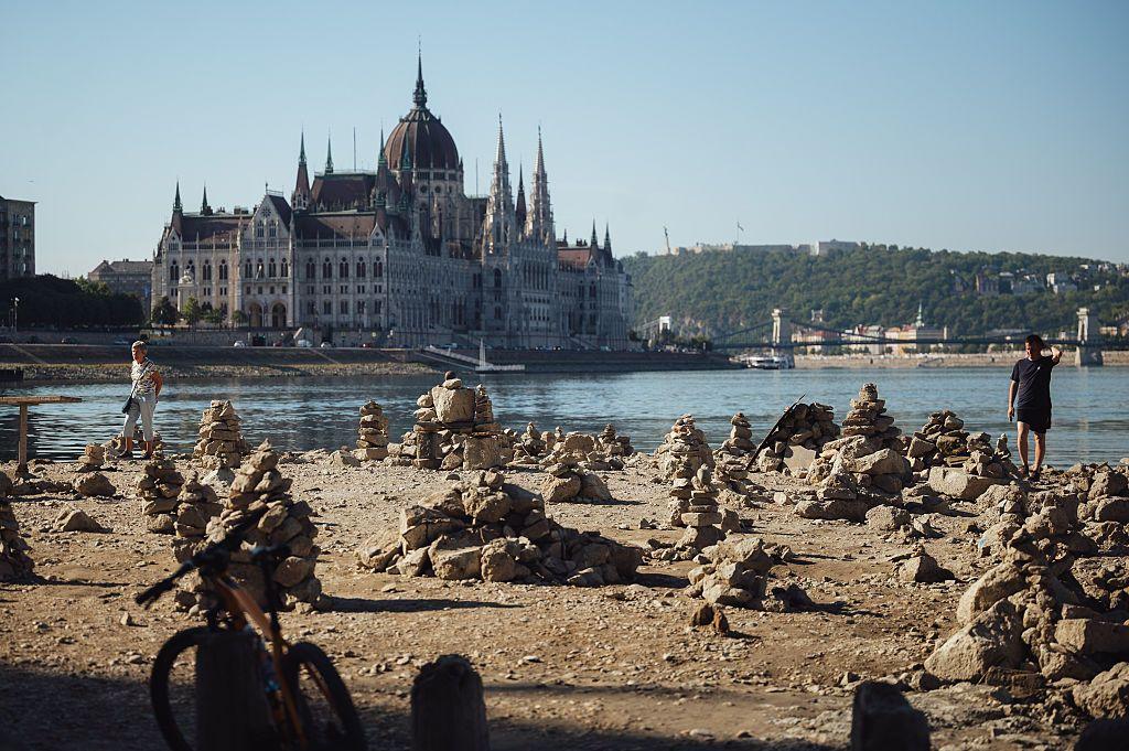 Una bicicleta y una persona en la parte del lecho del Danubio que la sequía ha hecho visible a su paso por Budapest. Más lejos, se aprecia el agua del río y un edificio histórico con una cúpula y varios pináculos bajo un cielo soleado.