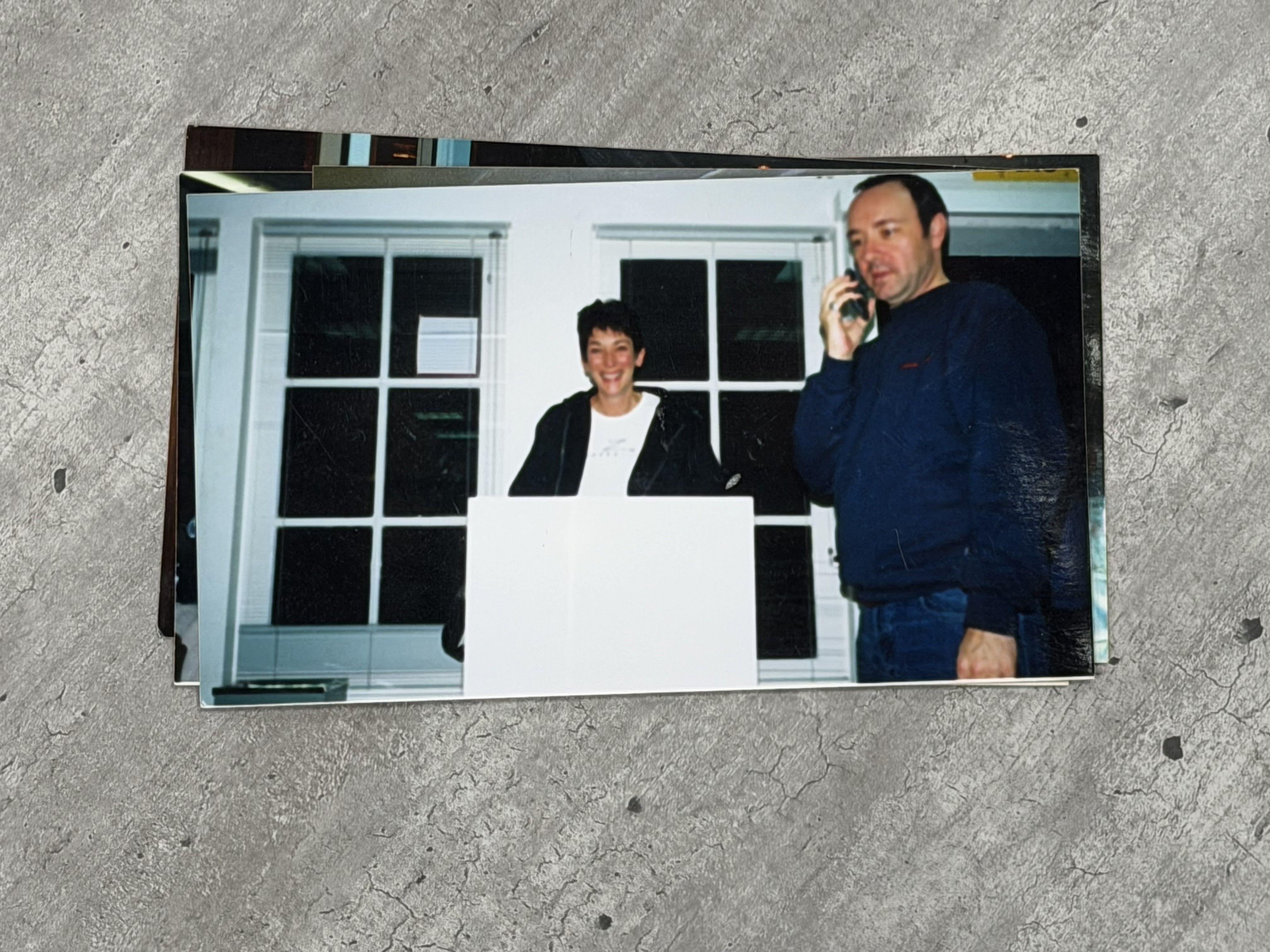 Una mujer, Ghislaine Maxwell, con el pelo corto, sonríe y está de pie detrás de un podio blanco. A su izquierda, un hombre, Kevin Spacey, lleva un teléfono móvil en la oreja derecha. Viste una camisa azul marino y vaqueros azules.