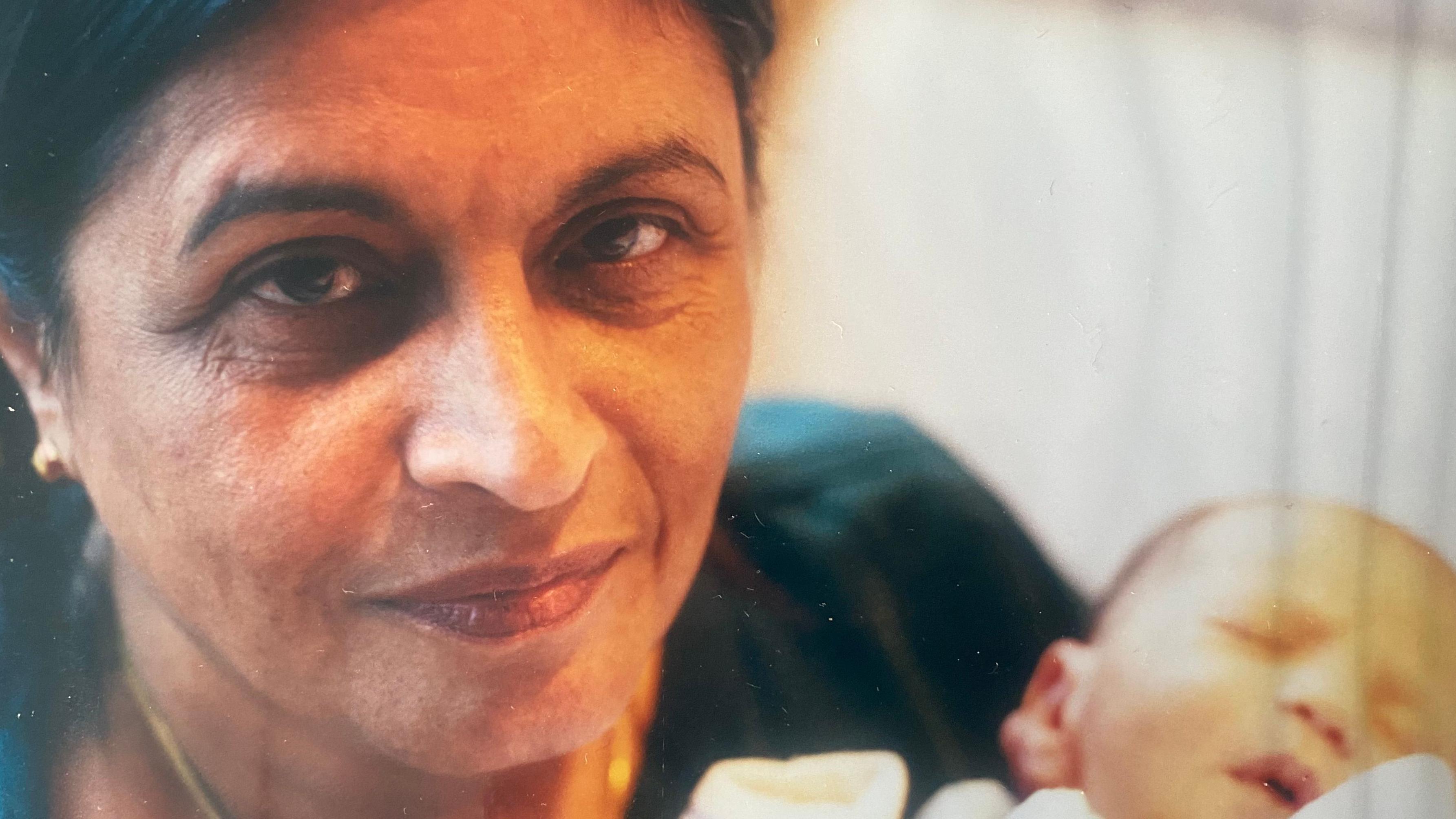 Samina Ali's mother cradles sleeping baby Ishmael in her arms while looking directly at the camera with a slight smile.