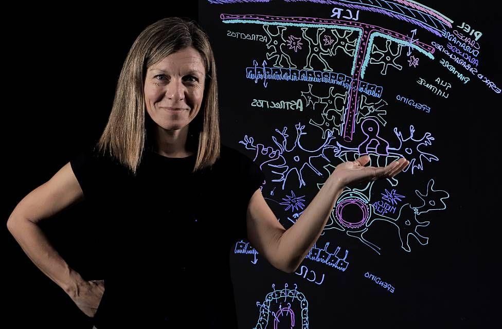 La profesora Noelia Valle, con una camiseta negra y pelo rubio en melena, frente a una pizarra con dibujos con tizas de colores sobre biología