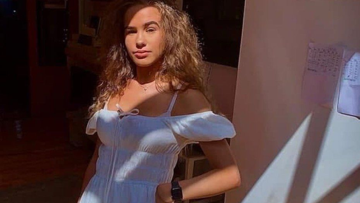 Brooke George is looking at the camera wearing a cotton off-the-shoulder dress. She has long wavy blonde hair and is posing for the camera with her hand on her hip