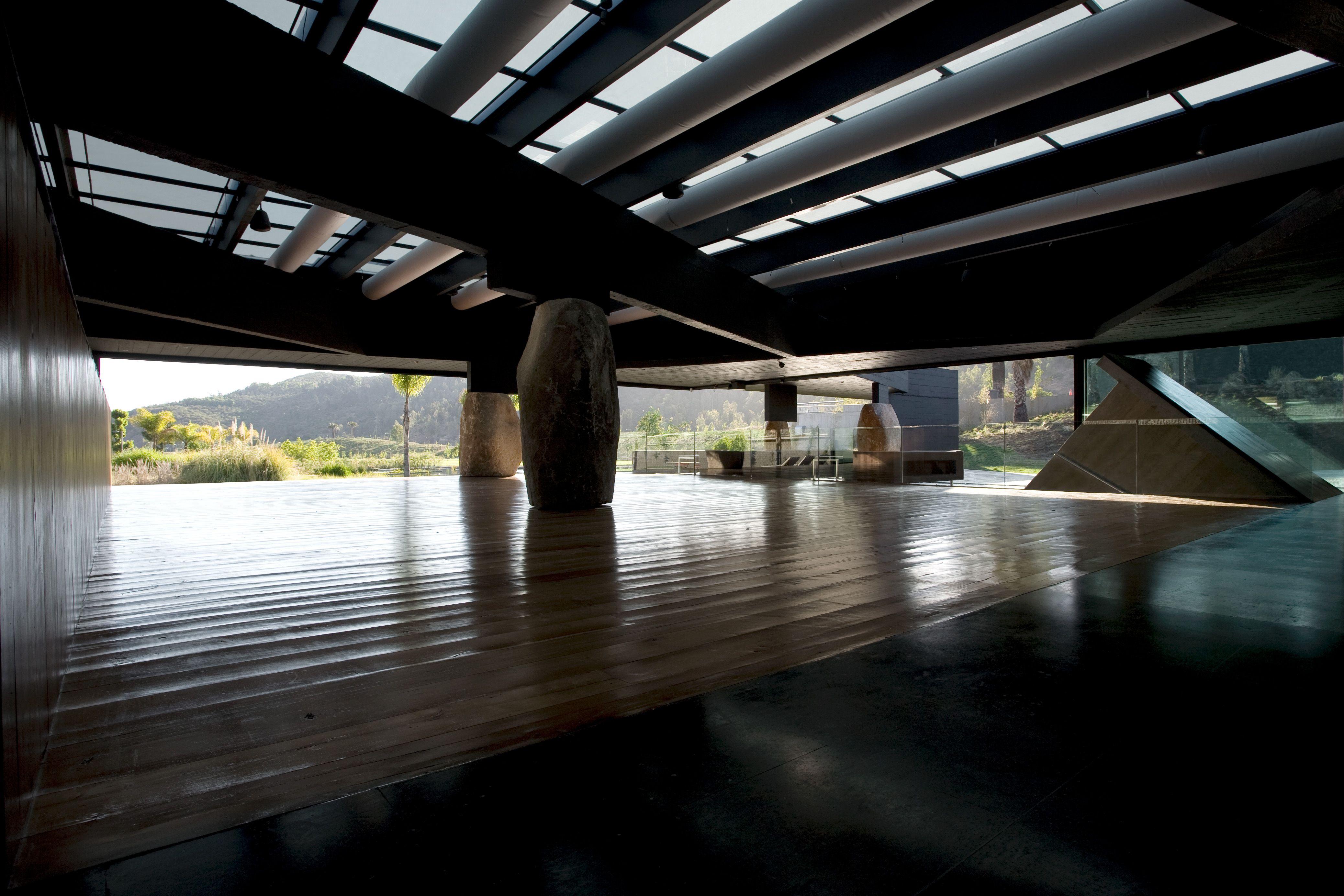 Interior sombreado con techo estructural visible, grandes vigas y columnas de piedra, abierto hacia un paisaje exterior iluminado.