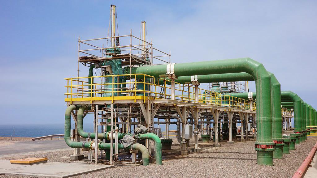 Redovi paralelnih zelenih cevi protežu se od podignute metalne platforme do popločanog tla ispod; more se vidi odmah iza.