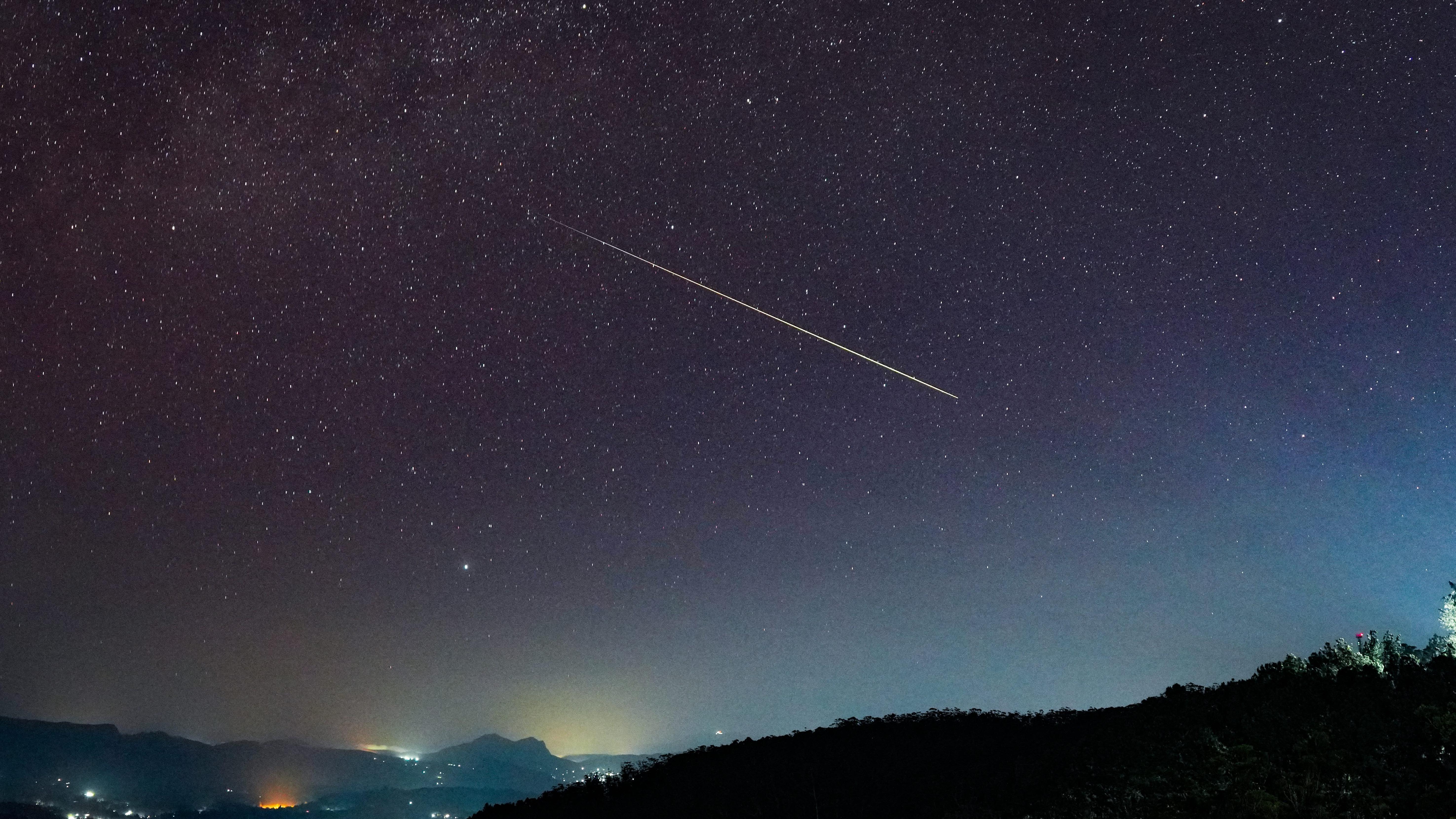 Veliki Perseid meteor dijagonalno prelazi zvezdano noćno nebo iznad Haputala, Šri Lanka - u prvom planu se vide tamna brda sa svetlima zgrada