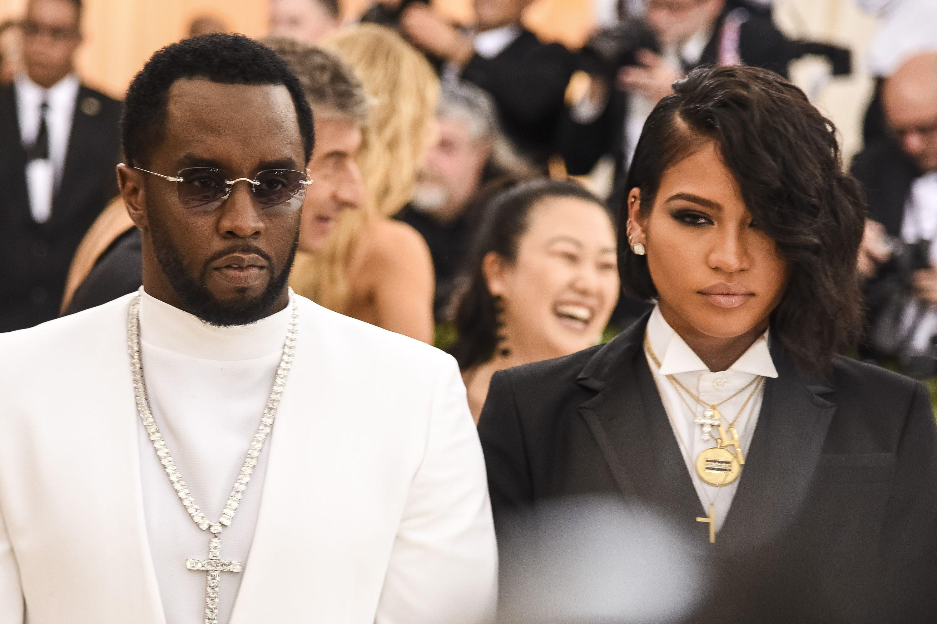 Sean Combs e ?Casandra Ventura, em trajes formais. Combs de terno branco e um crucifixo ornamentado e Ventura com terno preto e diversos colares, que incluem um crucifixo e um pendente circular