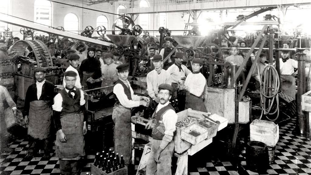 A imagem em preto e branco mostra o interior de uma f&aacute;brica no in&iacute;cio do s&eacute;culo 20, com dezenas de trabalhadores distribu&iacute;dos entre grandes m&aacute;quinas industriais. Muitos usam aventais e bon&eacute;s, enquanto operam equipamentos ou posam para a fotografia. O ambiente &eacute; amplo, iluminado por janelas altas, e transmite o ritmo intenso da produ&ccedil;&atilde;o industrial da &eacute;poca.