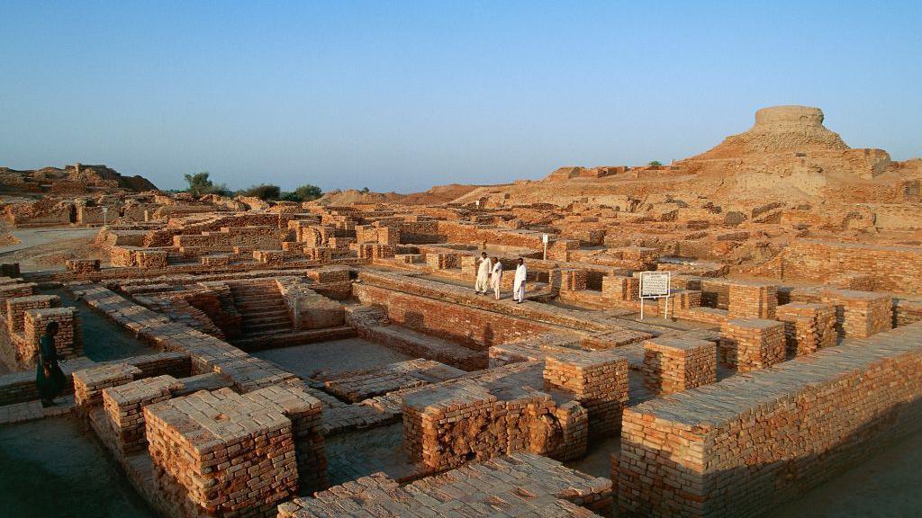 Uma vista elevada de ru&iacute;nas de tijolo, com funda&ccedil;&otilde;es retangulares e estruturas feitas de tijolos empilhados, em uma paisagem seca e arenosa. &Eacute; um dia ensolarado, com c&eacute;u azul