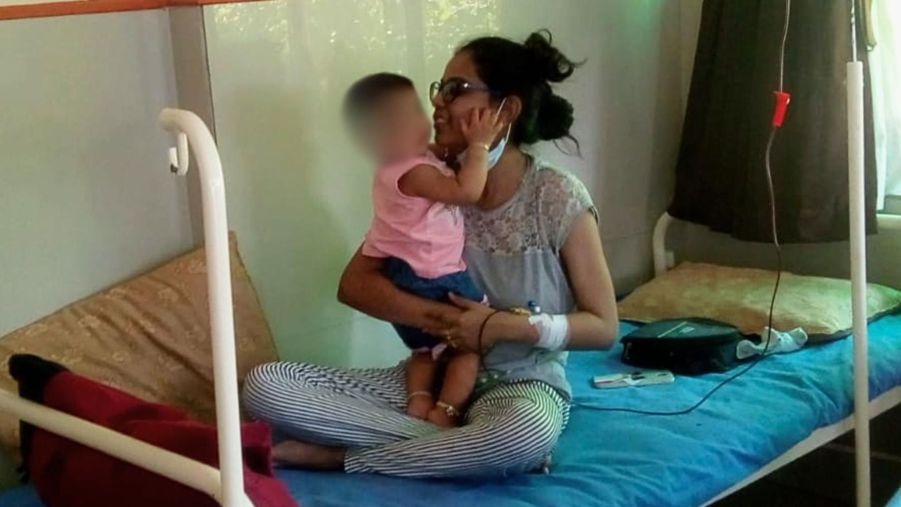 Kinjal is shown smiling sitting cross-legged on a metal hospital bed with a tube attached to her arm for a blood transfusion. Her baby daughter stands up in her lap
