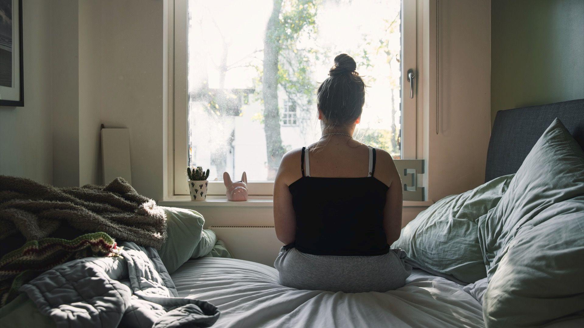 Rear view of a teenage girl looking out of the window whilst sitting on her bed