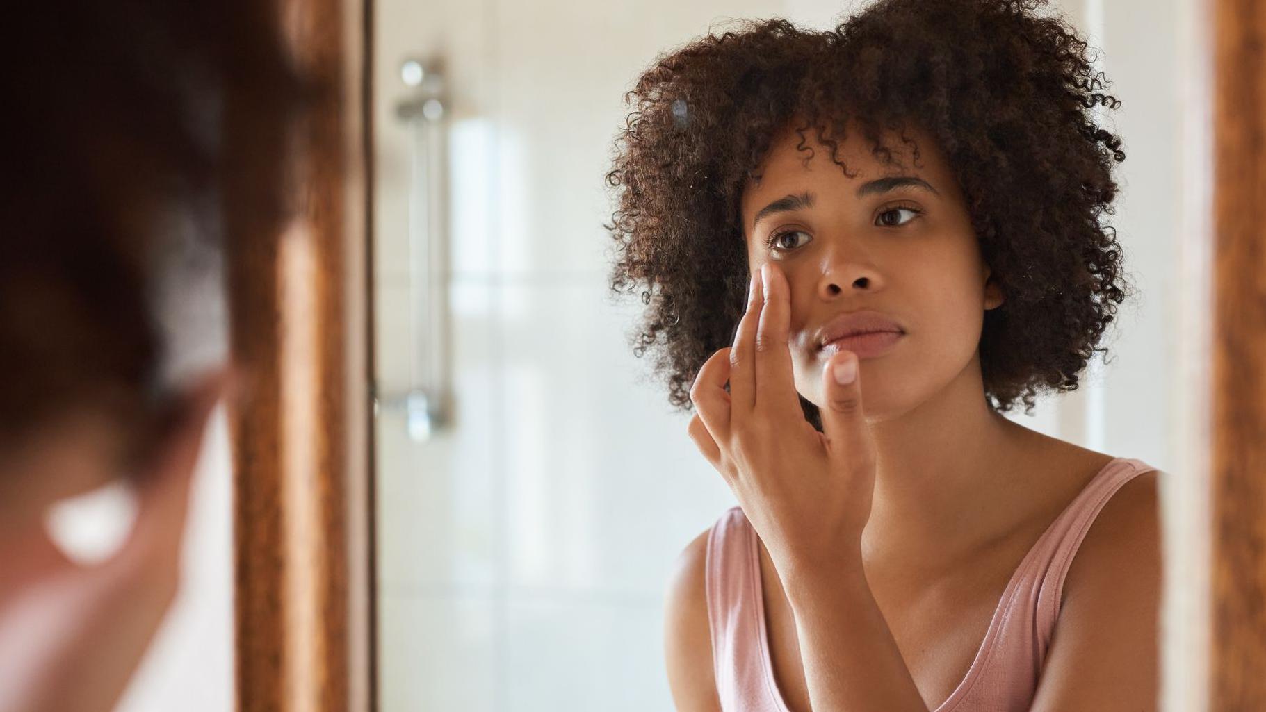 Una joven de cabello oscuro se mira en el espejo, tocándose la piel alrededor de los ojos con expresión sombría.