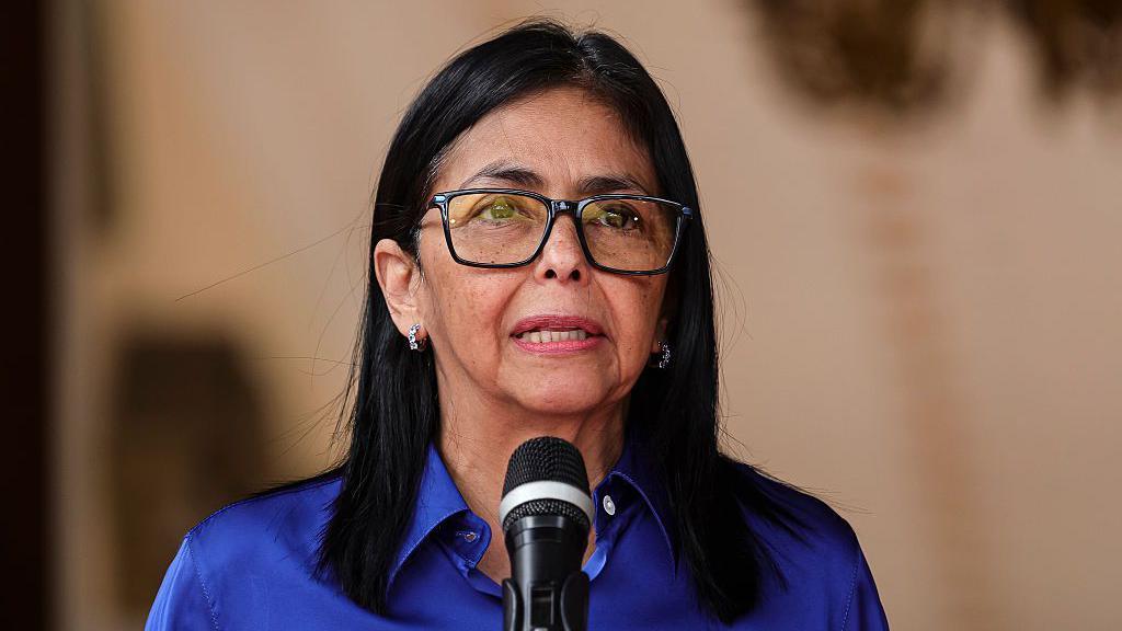 Delcy Rodríguez vestida con camisa azul hablando ante un micrófono. 