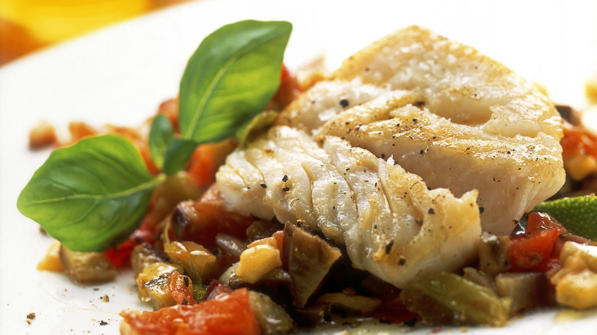 Um pedaço de bacalhau servido com legumes e decorado com uma folhinha verde de tempero