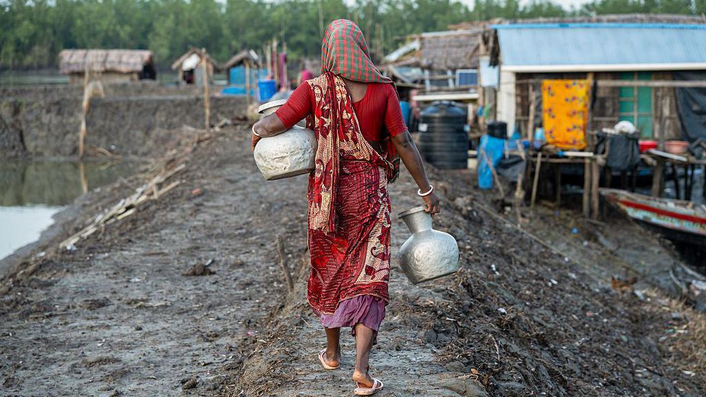 Una mujer con un vestido rojo y un recipiente en busca de agua potable.