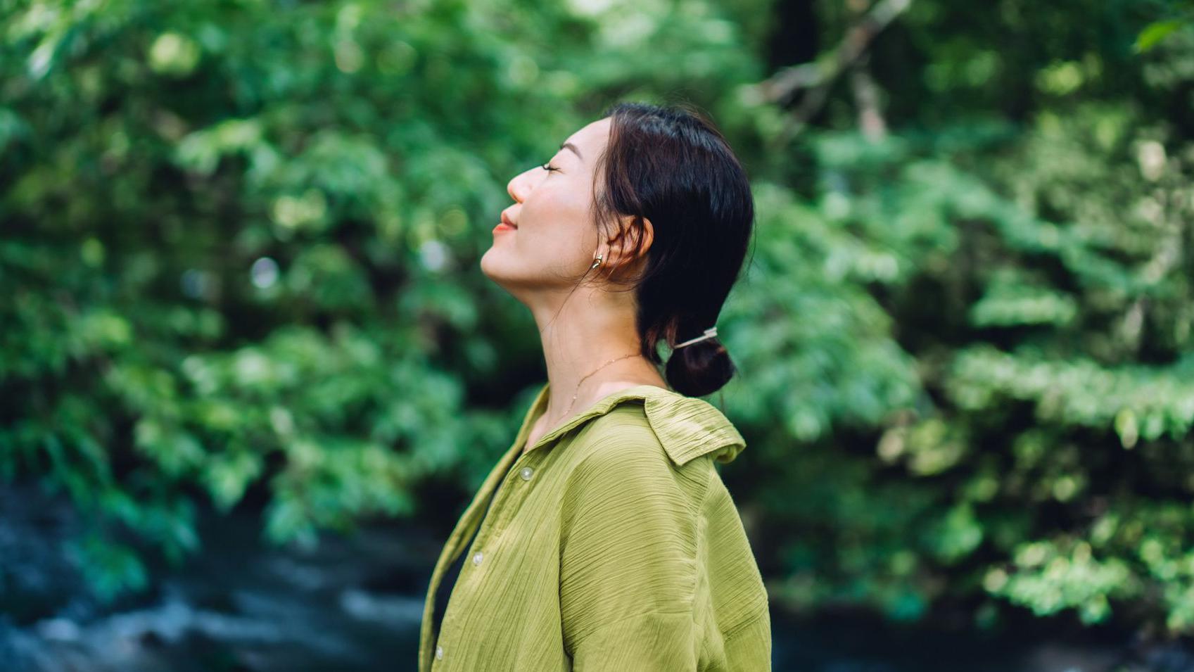 一位留著黑色長髮並紮成低馬尾的亞洲女性閉著眼睛仰望天空。她穿著一件綠色襯衫，背景是綠色的樹木。