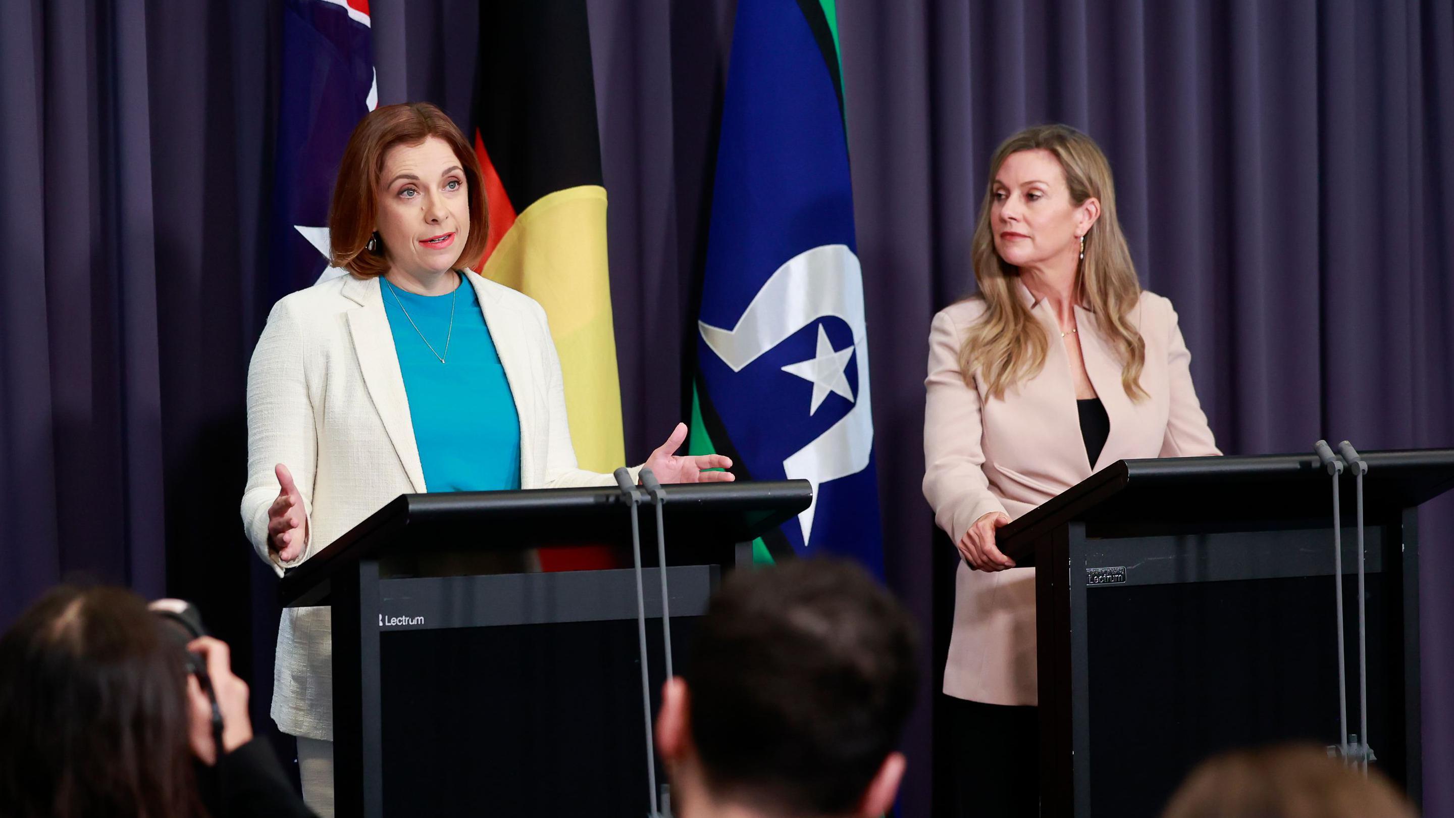 Anika Wells, vestida con chaqueta blanca, habla en un atril mientras una mujer rubia observa.