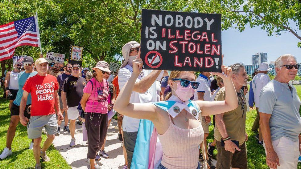 Manifestantes y una joven en primer plano con una pancarta que dice: "Nadie es ilegal en una tierra robada"