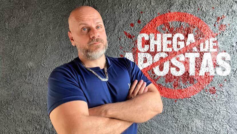 Rafael Santos, um homem corpulento, careca, de barba grisalha e olhos azuis, com os braços cruzados e uma expressão séria no rosto, a frente de um logotipo onde se lê: 